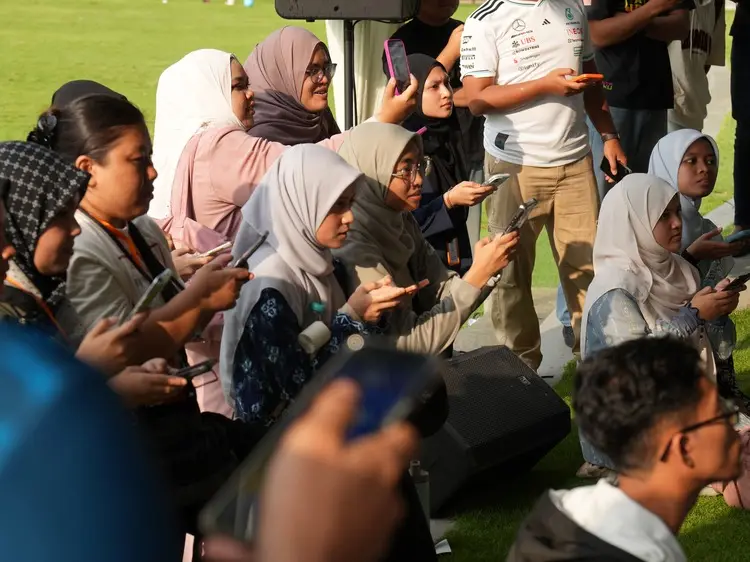 Sekumpulan pengunjung duduk di atas rumput menghadap pentas sambil merakam dengan telefon.