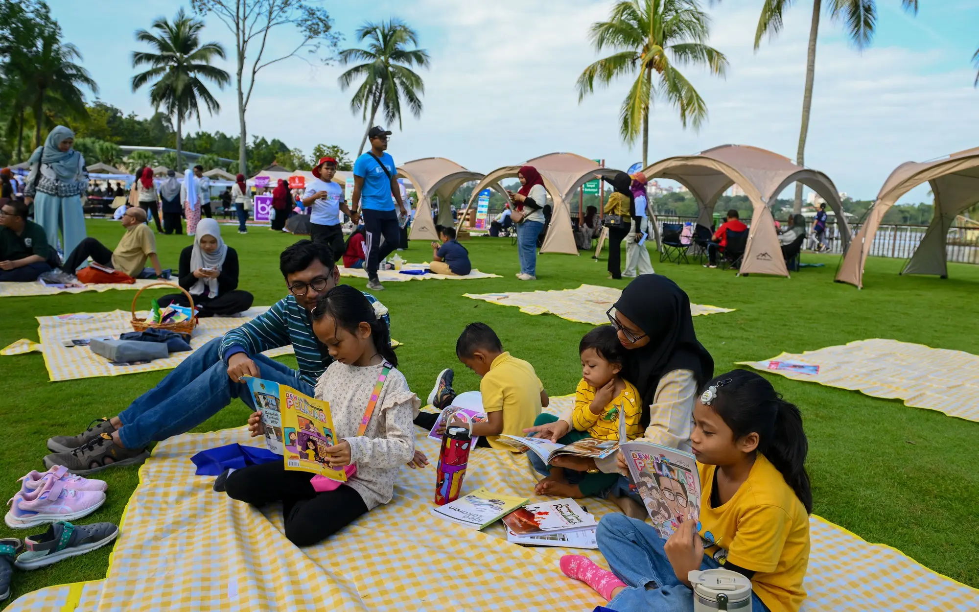 Beberapa keluarga duduk di atas tikar berkelah di atas rumput sambil membaca buku bergambar, dengan khemah dan tasik di latar belakang.